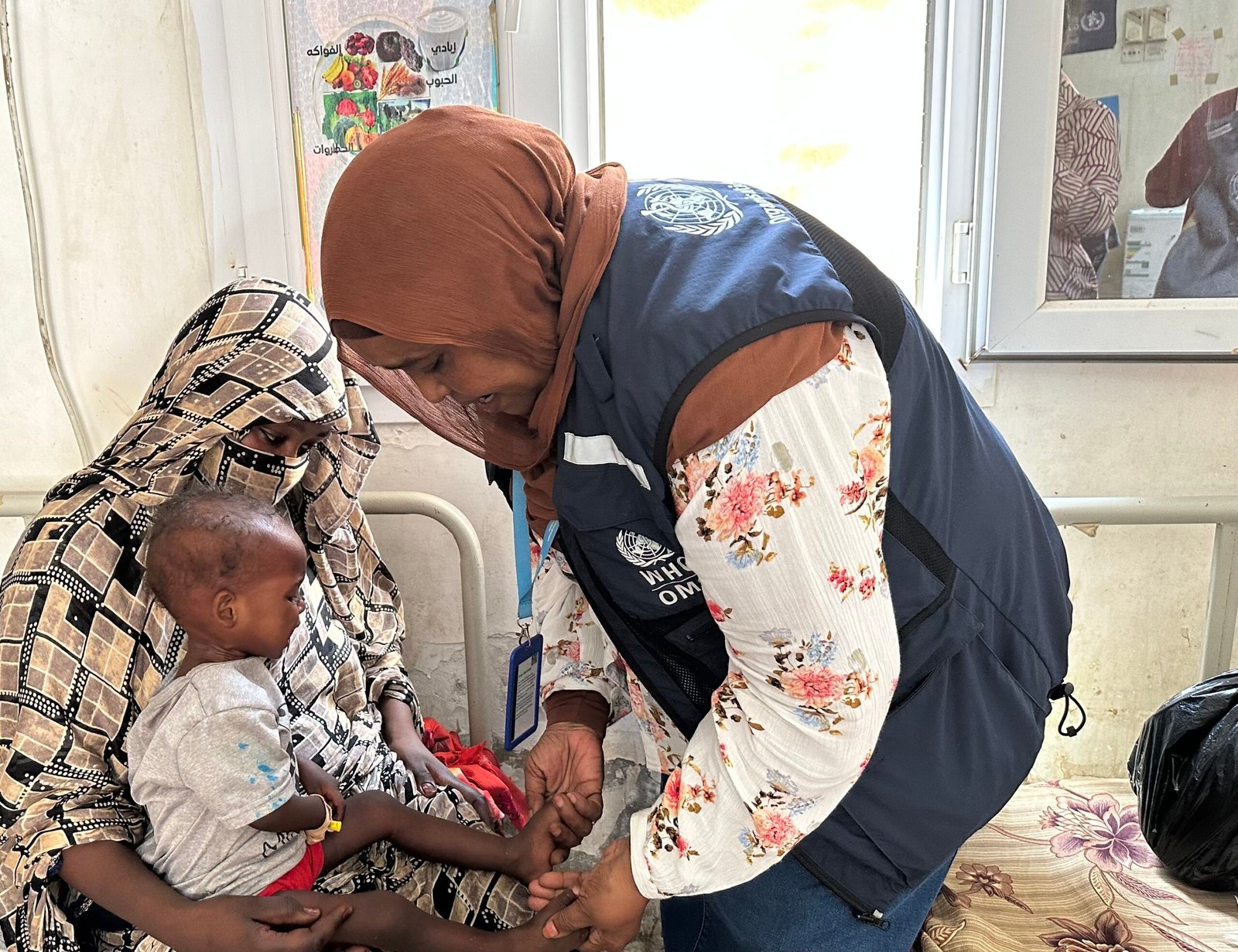 عاملة صحية تفحص طفلاً بين ذراعي والدته في مركز علاج سوء التغذية ببورتسودان.
